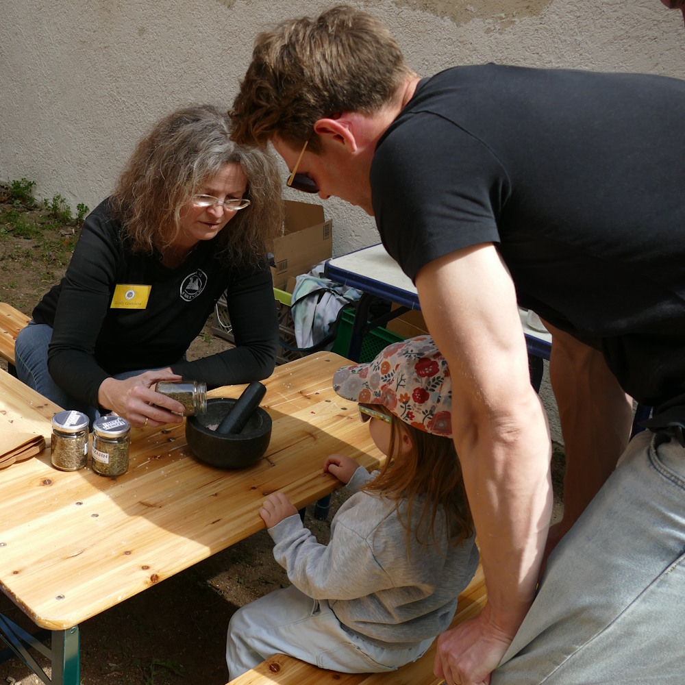 Eine Frau erklärt einem Vater und einem kleinen Kind an einem Holztisch das Zermahlen von Kräutern im Mörser während eines Mitmachangebots auf der Strahlenburg.